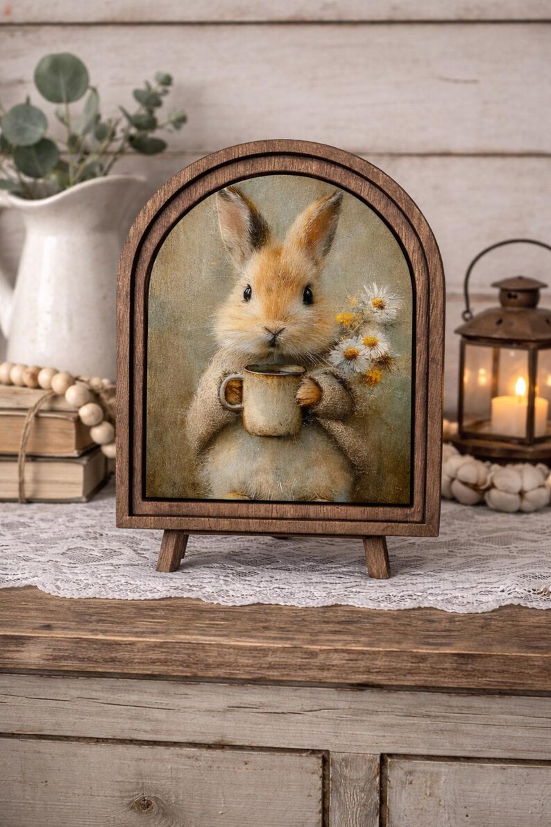 May include: A wooden framed print of a bunny holding a mug and flowers. The bunny is light brown with long ears and is holding a small brown mug. The print is displayed on a small wooden easel.