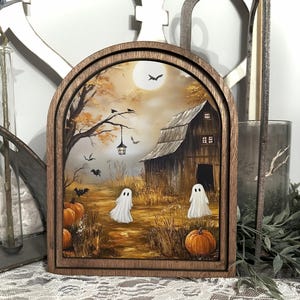 May include: A Halloween-themed painting in a wooden frame. The artwork depicts a spooky scene with a barn, pumpkins, ghosts, and bats under a full moon. The color palette includes shades of brown, orange, and white, creating a festive autumn atmosphere.