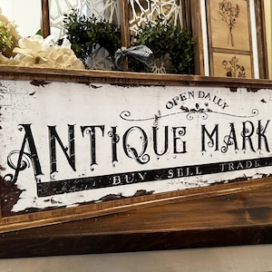 Op de afbeelding: Een rustiek houten bord met witte verf en zwarte letters met de tekst "Open Daily Antique Market Buy Sell Trade". Het bord heeft een verweerde uitstraling en is perfect voor een winkel of voor thuisdecoratie.