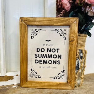 Może przedstawiać: Oprawiony znak z napisem "PLEASE DO NOT SUMMON DEMONS in the bathroom". Rama jest z jasnego drewna. Czarne ozdoby i grafika nietoperza u góry. Znak znajduje się na drewnianej powierzchni.