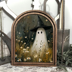 Peut inclure: Une œuvre d'art encadrée représentant un fantôme portant un chapeau de sorcière noir, entouré de fleurs sauvages. Le fantôme est blanc avec des yeux noirs, et l'œuvre est encadrée dans une arche en bois foncé. L'arrière-plan est vert foncé et marron.