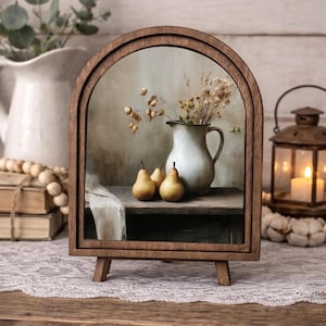 May include: A wooden framed still life featuring a white pitcher with dried flowers, three pears, and a small table. The frame has an arched top and is supported by small wooden legs. A candle lantern is in the background.