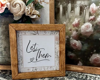 Op de afbeelding: Een ingelijst houten bord met een witte achtergrond en een bloemenpatroon. Het bord zegt "Let Them" met een klein hartje.