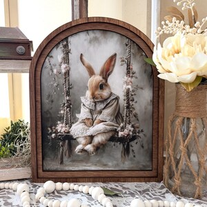 May include: A framed print featuring a rabbit on a swing, adorned with floral accents. The artwork is set within a dark brown, arched wooden frame. The rabbit is dressed in a vintage-style dress. The overall aesthetic is soft and whimsical.