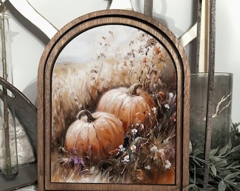 May include: An arched wooden frame containing a printed image of two orange pumpkins nestled in a field of dried grasses and small white flowers. The frame features a dark brown finish with a smooth, rounded top edge and a layered border design.