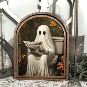 May include: A wooden framed art piece featuring a white ghost sitting on a toilet, reading a newspaper. The ghost has black oval eyes and is draped in a white sheet. The background includes a green wall, orange flowers, and a toilet.