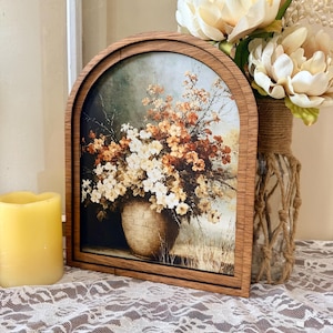 Può includere: Una cornice in legno ad arco contenente una stampa di una composizione floreale in un cesto intrecciato. Il bouquet presenta fiori bianchi e arancioni su uno sfondo tenue. La cornice ha una finitura in legno naturale e un bordo superiore arrotondato.