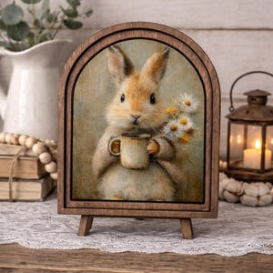 May include: A wooden framed print featuring a charming illustration of a bunny holding a mug and daisies. The bunny is light brown with a cream-colored belly. The frame is arched at the top and supported by a small easel.