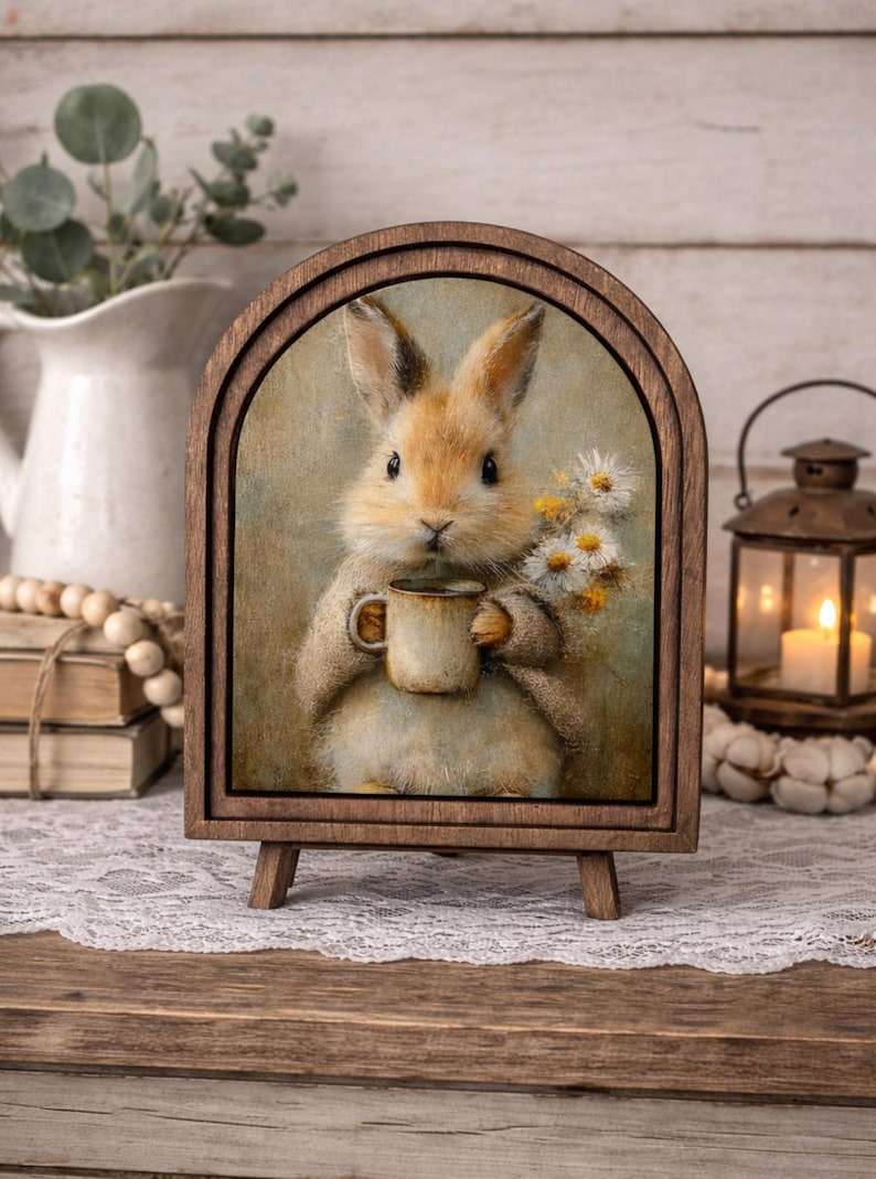 May include: A wooden framed print featuring a charming illustration of a bunny holding a mug and daisies. The bunny is light brown with a cream-colored belly. The frame is arched at the top and supported by a small easel.