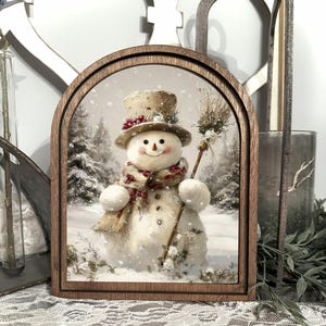 May include: A framed winter scene features a smiling snowman wearing a top hat, scarf, and holding a broom. The snowman is set against a snowy forest backdrop. The frame is made of dark wood. A charming holiday decoration.
