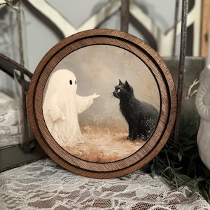 Op de afbeelding: Rond houten kunstwerk met een witte geest en een zwarte kat. De geest, met twee zwarte ogen, wijst naar de kat. De kat zit met opgerolde staart en kijkt naar de geest. De achtergrond is een gedempt, neutraal landschap.