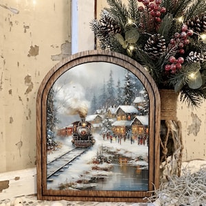 May include: An arched wooden frame containing a winter scene print of a steam locomotive traveling on tracks through a snow-covered village with pine trees, glowing windows, and a small stream reflecting the light under a cloudy sky.