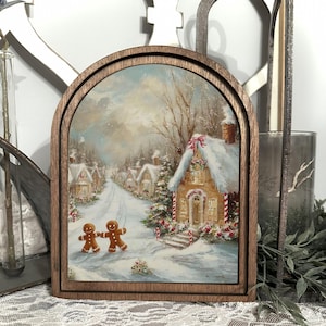 May include: Wooden framed art print featuring a winter scene with snow-covered houses and a road. Two gingerbread men are in the foreground. The frame has a dark brown finish and an arched top. The artwork has a warm, inviting color palette.