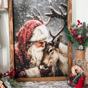 May include: A framed print shows Santa Claus hugging a reindeer in a snowy setting. Santa is in a red suit and hat with white fur. The reindeer has large antlers. The frame is dark wood. The image has a Christmas theme.