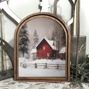 Könnte beinhalten: Ein gerahmter Druck mit einer Winterszene mit einer roten Scheune unter einem schneebedeckten Himmel. Das Kunstwerk befindet sich in einem holzfarbenen, bogenförmigen Rahmen, der eine rustikale Ästhetik erzeugt. Der Druck zeigt eine schneebedeckte Landschaft mit Bäumen und einem Holzzaun.