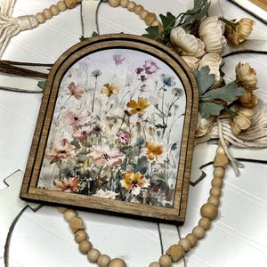 May include: A framed watercolour painting of a field of wildflowers in shades of pink, yellow, and white. The flowers are arranged in a loose, natural style, and the background is a soft, muted blue.
