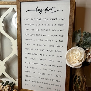 Op de afbeelding: Een ingelijst bord met een witte achtergrond en zwarte tekst die "buy dirt" luidt. De tekst gaat verder met een lijst van levenslessen, zoals "Vind degene zonder wie je niet kunt leven" en "Je kunt geluk niet kopen, maar je kunt aarde kopen".