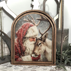 May include: A wooden framed print featuring Santa Claus embracing a reindeer in a snowy scene. Santa is wearing a red suit and hat with white fur trim. The reindeer has large antlers and is brown and white.