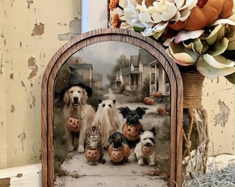 May include: An arched wooden frame containing a print of a golden retriever wearing a witch hat, a cat, a ghost-costumed animal, a dachshund, a bat-winged dog, and a small dog, all holding miniature jack-o'-lantern buckets on a neighborhood street.
