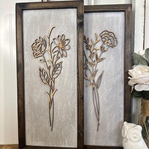 May include: Two wooden framed wall art pieces with a distressed white background. Each piece features a different floral design cut out of wood and painted gold.