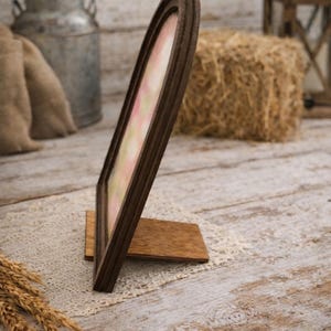 May include: A brown wooden picture frame with an arched top, standing on a wooden base. The frame holds a pastel-colored image. The background includes a hay bale, a metal container, and a rustic wooden surface.