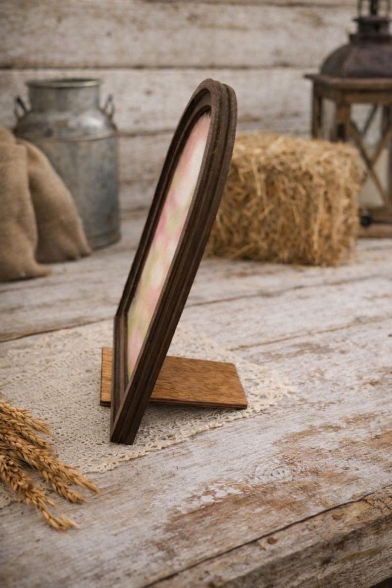 May include: A brown wooden picture frame with an arched top, standing on a wooden base. The frame holds a pastel-colored image. The background includes a hay bale, a metal container, and a rustic wooden surface.