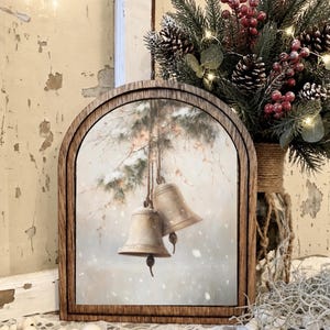May include: An arched wooden frame containing a print of two hanging metallic bells suspended from a snow-covered pine branch. The artwork features a soft, muted color palette with white snowflakes falling against a light, neutral background.