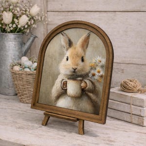 May include: A framed print of a bunny holding a mug, with a bouquet of daisies. The artwork is displayed on a small wooden easel. The frame is arched at the top and made of dark wood. The background is a neutral color.