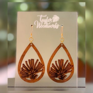 May include: A pair of wooden earrings with a teardrop shape and a laser-cut design of a flower. The earrings are brown and have gold hooks.