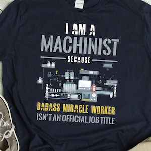 May include: A navy blue t-shirt with the text "I am a Machinist because badass miracle worker isn't an official job title" in white and yellow lettering. The shirt features a graphic of a factory assembly line with workers.