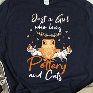 Può includere: Una maglietta blu navy con il testo "Just a Girl who loves Pottery and Cats". Il design presenta due gatti calico che fiancheggiano un vaso di ceramica con un motivo floreale.