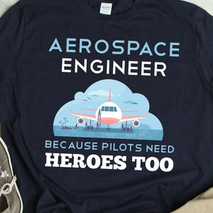 May include: A navy blue t-shirt with the text "Aerospace Engineer" and "Because pilots need heroes too". The shirt features a cartoon illustration of an airplane being worked on by mechanics.