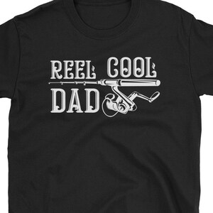May include: Black t-shirt with the text "REEL COOL DAD" in white, featuring a fishing rod and reel graphic. The design is centered on the front of the shirt. The shirt is made of a soft material.