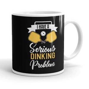 May include: White coffee mug with a black background and a yellow and black pickleball paddle design. The text on the mug reads "I have a serious dinking problem."