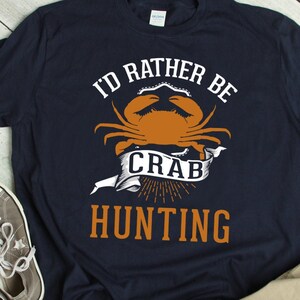 May include: A navy blue t-shirt with the text "I'd rather be crab hunting" in white and orange. A cartoon crab is featured in the design.