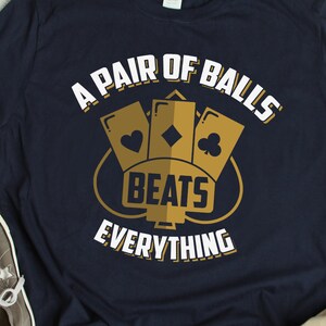May include: A navy blue t-shirt with a white graphic design. The graphic features three playing cards with the text "A Pair of Balls Beats Everything".