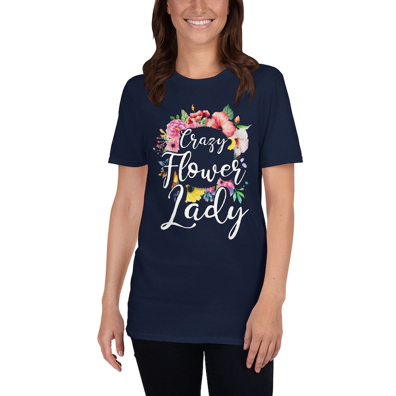 May include: Navy blue t-shirt with the text "Crazy Flower Lady" in white script, surrounded by a floral wreath of pink, red, and yellow flowers. The shirt is a crew neck and short-sleeved.