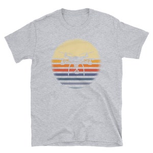 May include: A gray t-shirt with a vintage-style graphic of a drone flying in front of a sunset. The sunset is depicted with orange, yellow, red, and blue stripes.