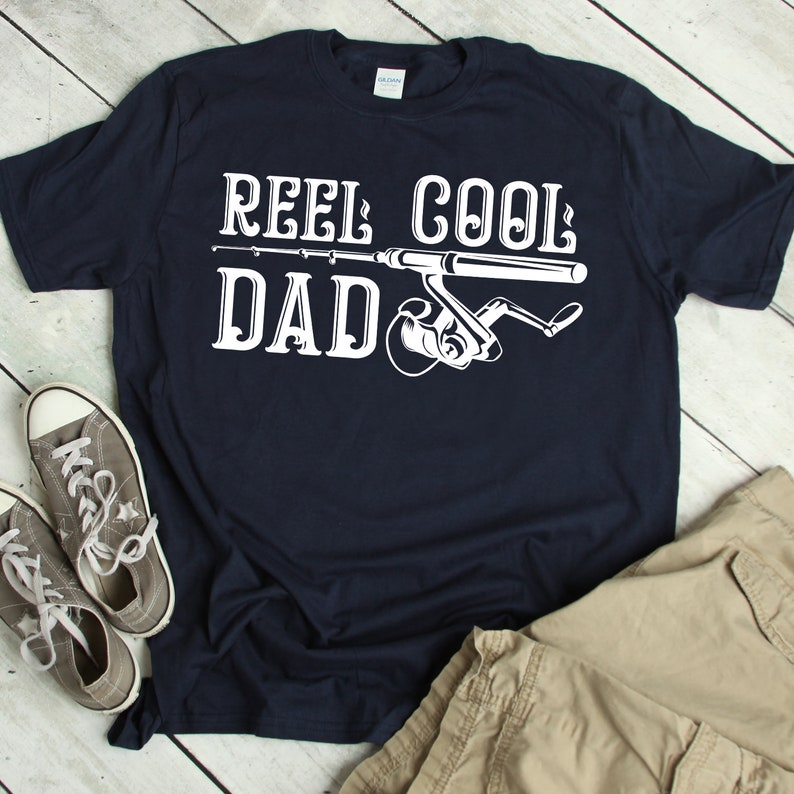 May include: Navy blue t-shirt with the text "REEL COOL DAD" in white, accompanied by a fishing rod and reel graphic. Gray sneakers and khaki shorts are visible. A casual, comfortable style.