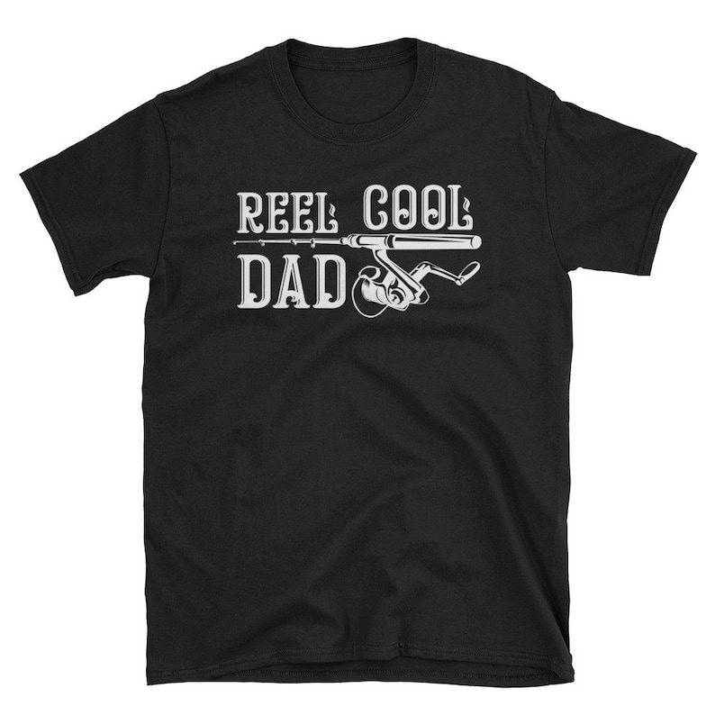 May include: Black t-shirt with the text "REEL COOL DAD" in white, featuring a fishing rod and reel graphic. The design is centered on the front of the shirt. The shirt is made of a soft material.