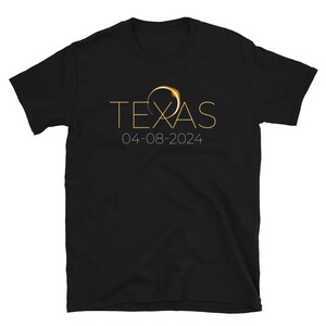 Puede incluir: Camiseta negra con un gráfico dorado de un anillo con un eclipse solar en su interior. El texto "TEXAS" está por encima del gráfico y la fecha "04-08-2024" está por debajo.