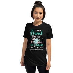 May include: Black t-shirt with a teal graphic that says "I am a Floral Designer not a magician but I can see why you are confused". The graphic is surrounded by small pink and blue flowers.