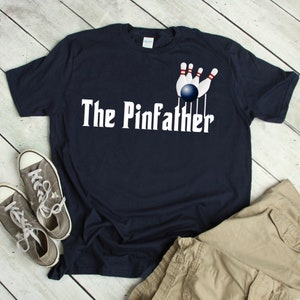 May include: A navy blue t-shirt with a white graphic of a bowling ball knocking down pins. The text "The Pinfather" is printed in white below the graphic.