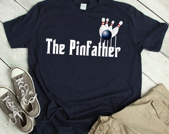 May include: A navy blue t-shirt with a white graphic of a bowling ball knocking down pins. The text "The Pinfather" is printed in white below the graphic.