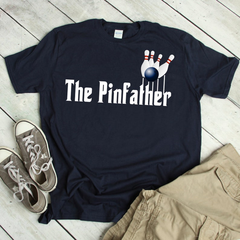 May include: A navy blue t-shirt with a white graphic of a bowling ball knocking down pins. The text "The Pinfather" is printed in white below the graphic.