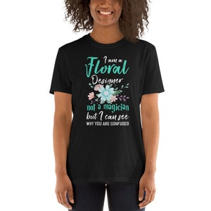May include: Black t-shirt featuring a teal green graphic that reads "I am a Floral Designer not a magician but I can see why you are confused". The graphic also includes a floral design with pink, blue, and purple flowers.