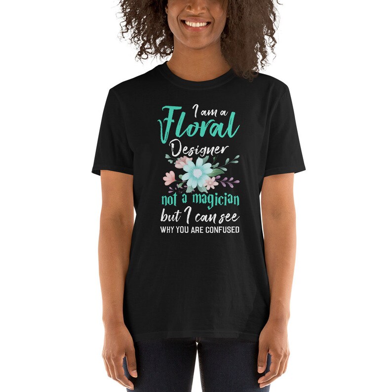 May include: Black t-shirt featuring a teal green graphic that reads "I am a Floral Designer not a magician but I can see why you are confused". The graphic also includes a floral design with pink, blue, and purple flowers.