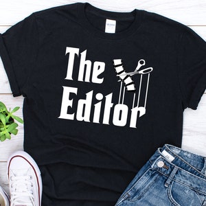 Könnte beinhalten: Schwarzes T-Shirt mit weißem Text "The Editor" und einer Grafik von Filmstreifen, die mit einer Schere geschnitten werden.
