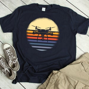 May include: A navy blue t-shirt with a vintage-style graphic of a drone flying in front of a sunset. The graphic is in a circle with a yellow background and colorful stripes.