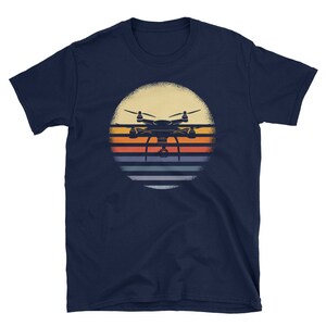 May include: A navy blue t-shirt with a vintage-style graphic of a drone flying over a sunset. The graphic features a silhouette of a drone with a camera, set against a background of orange, yellow, and blue stripes.